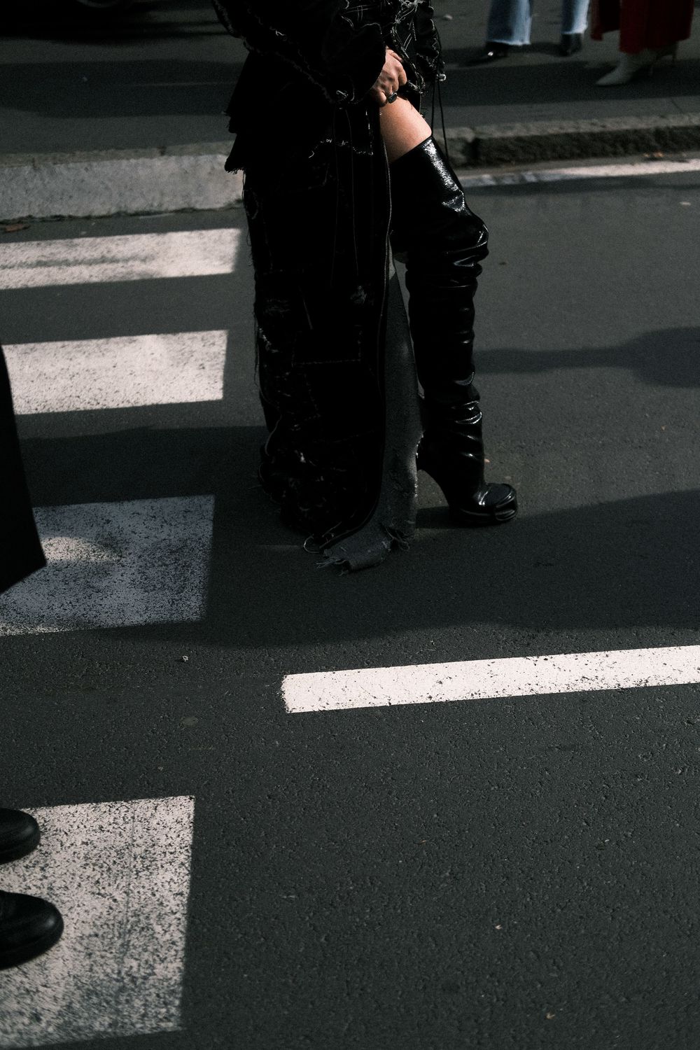 Legs in black boots standing on a crosswalk with dramatic shadows and white painted lines on dark pavement.