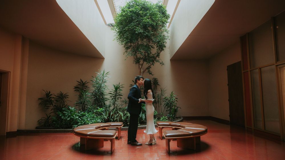 A panoramic view of an indoor hallway with ping pong tables and potted plants against beige walls.