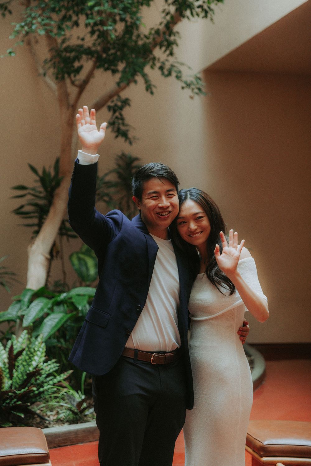 A couple in formal attire wave and smile together in front of an indoor garden setting.