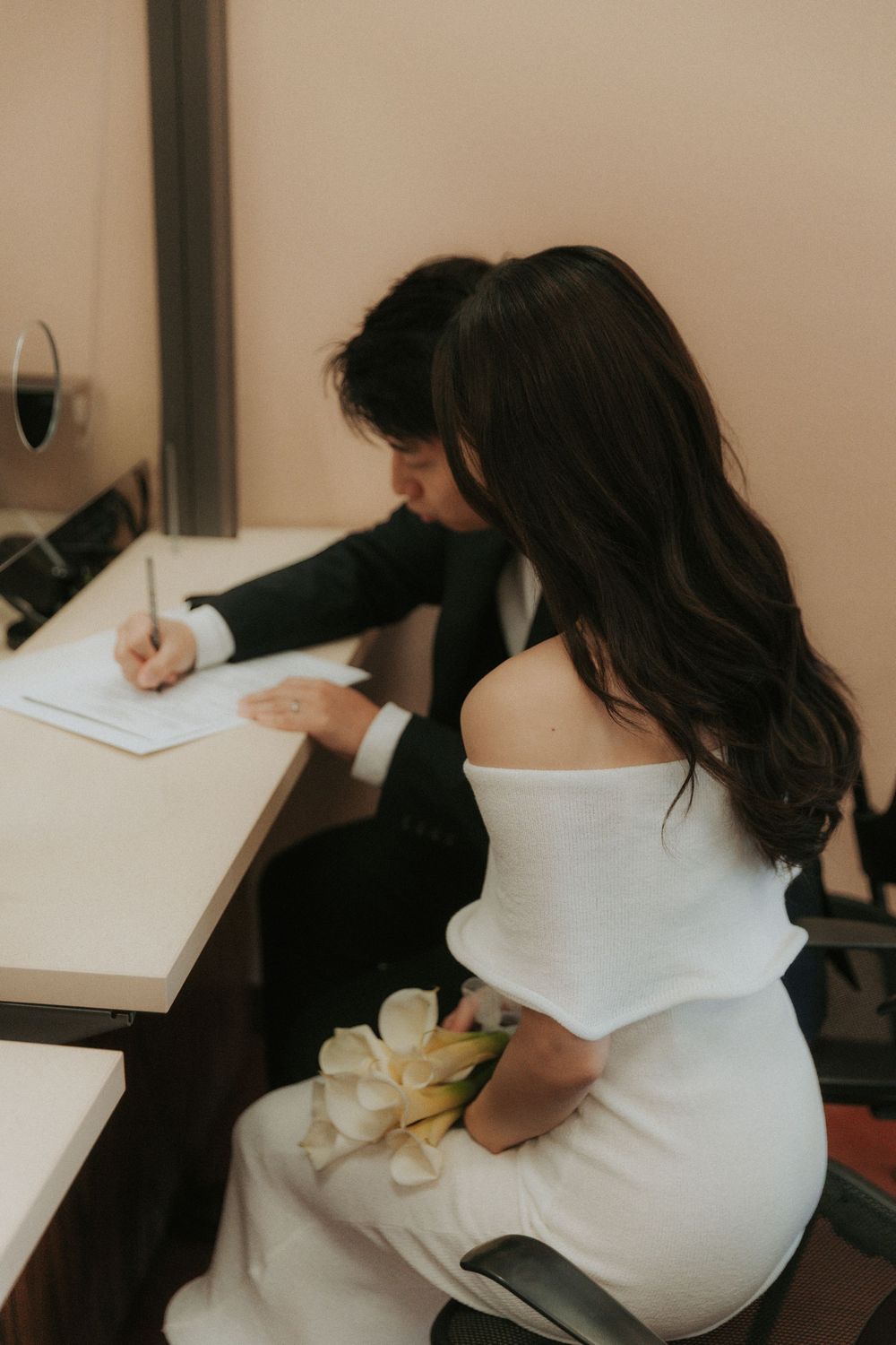 A couple signs documents together at a desk while the bride holds a white flower bouquet.