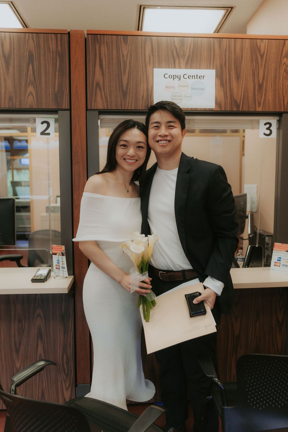 A couple in formal wedding attire stands in front of a copy center counter at a courthouse.