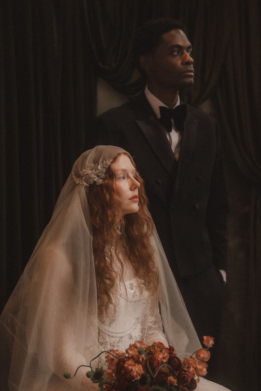 A bride in a white dress and veil holds an autumn bouquet while standing next to a groom in a black tuxedo.