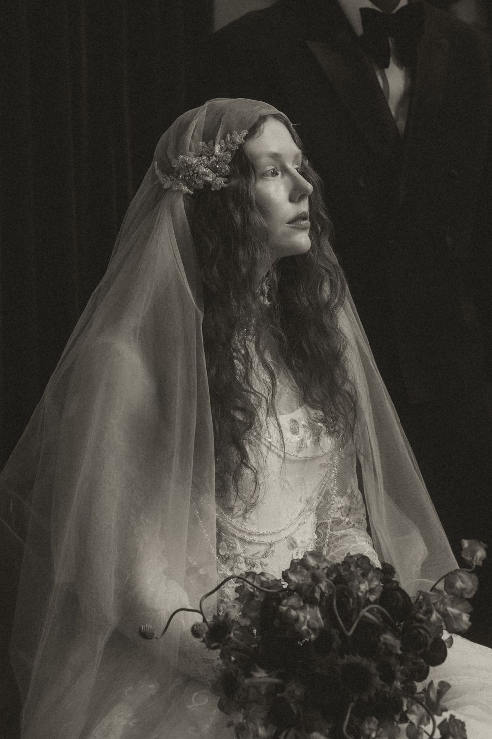 A vintage black and white photograph shows a bride in a delicate veil and floral crown holding a bouquet.