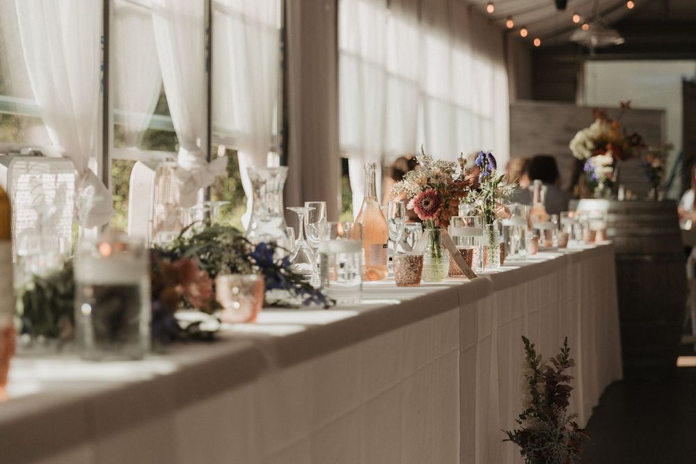Long wedding banquet table decorated with wildflower arrangements, candles, and glassware under string lights.