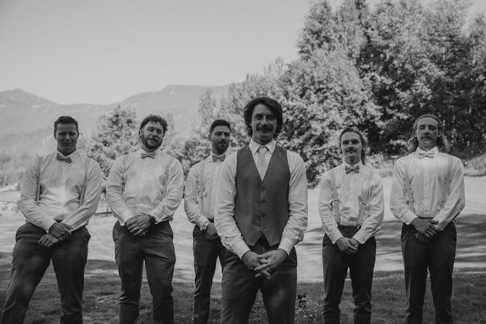 Black and white portrait of groom standing with groomsmen in white shirts against mountain backdrop.