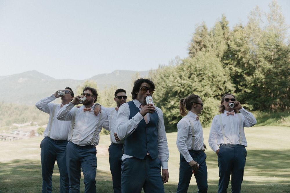 Groomsmen in blue pants and white shirts celebrating outdoors with mountains and trees in background.