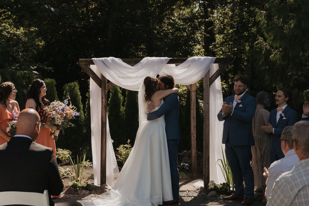 Bride and groom share first kiss under a white draped wooden arch at an outdoor garden wedding ceremony.