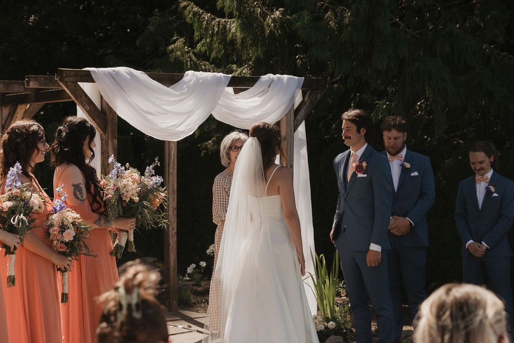 Bride in white gown and veil stands at altar with groomsmen in blue suits during outdoor wedding ceremony.