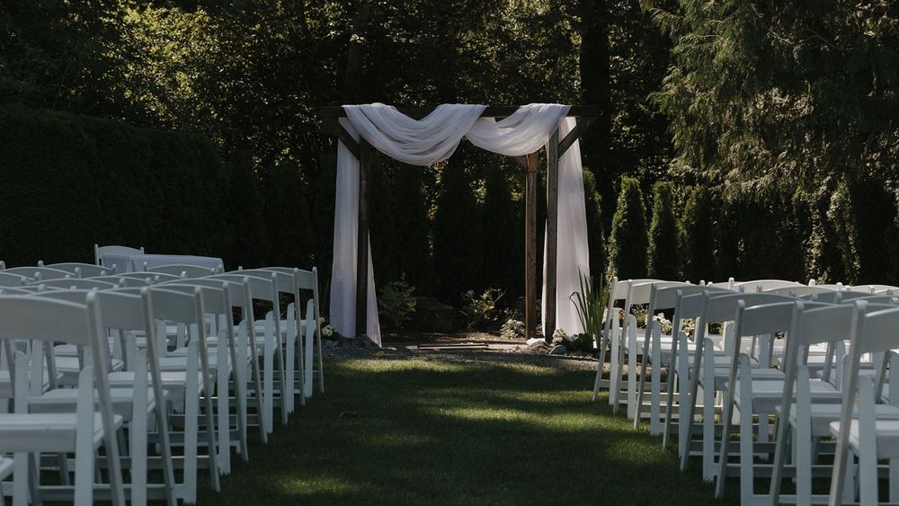 White folding chairs line a grass aisle leading to a white draped wooden wedding ceremony arch outdoors.