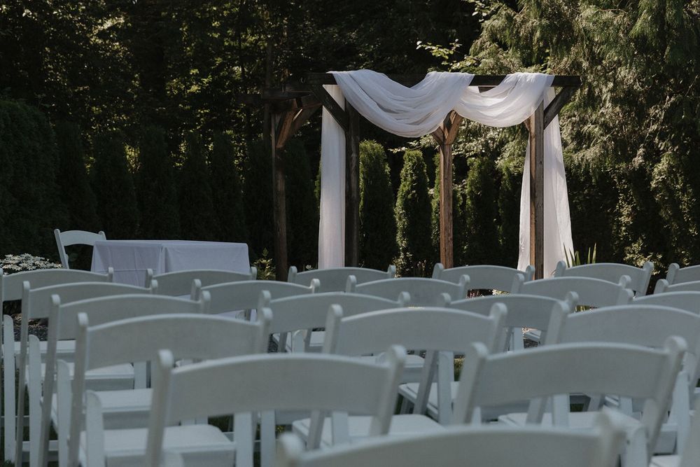 White folding chairs arranged before a draped wooden arch in a shaded outdoor wedding ceremony space.