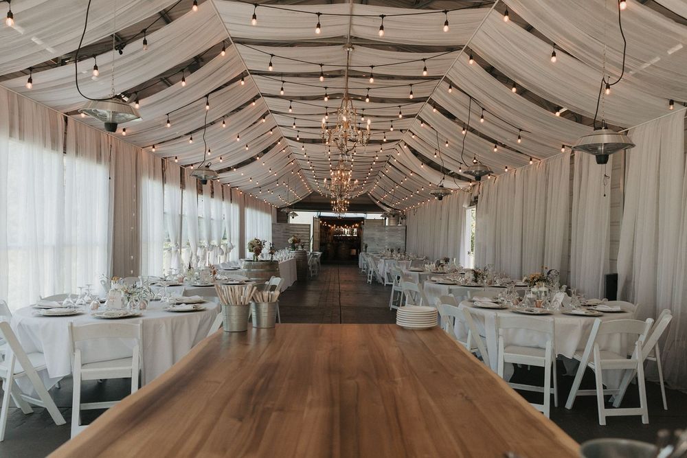 Elegant wedding reception hall with string lights, chandeliers, white draping, and long wooden dining tables.