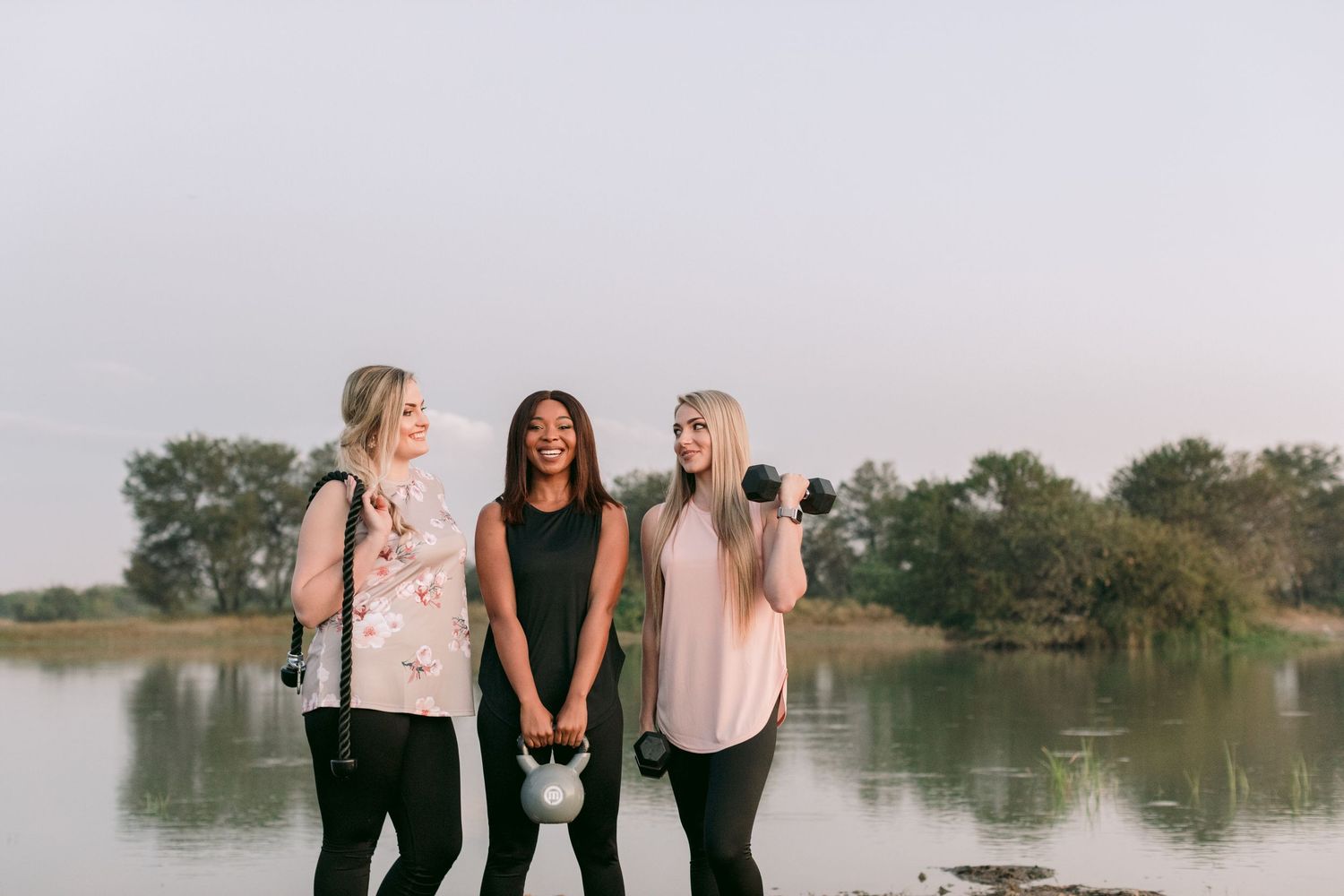 Three women laughing outdoors near a lake while holding kettlebells and dumbbells during a workout.