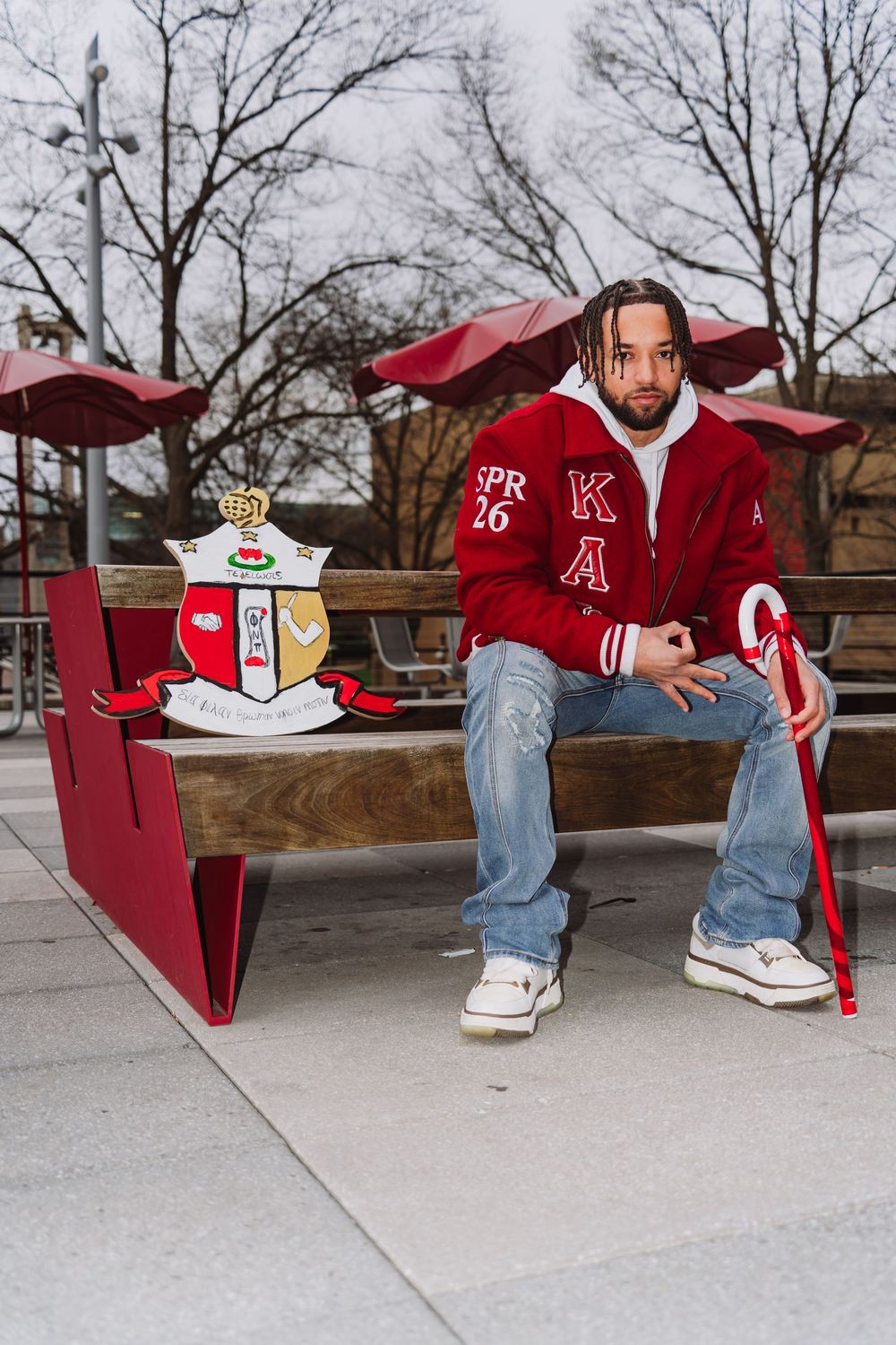 A person sits on a red outdoor bench wearing a varsity jacket, jeans and sneakers on a cloudy day.