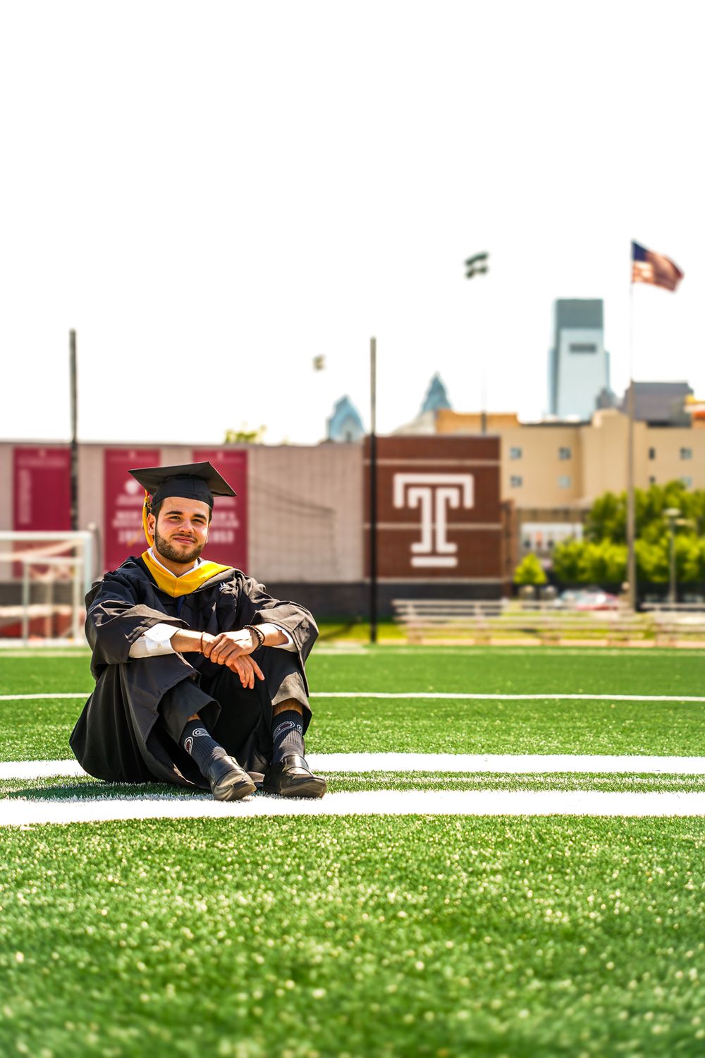 Graduate in cap and gown sits on football field with Temple University buildings and logo visible in background.