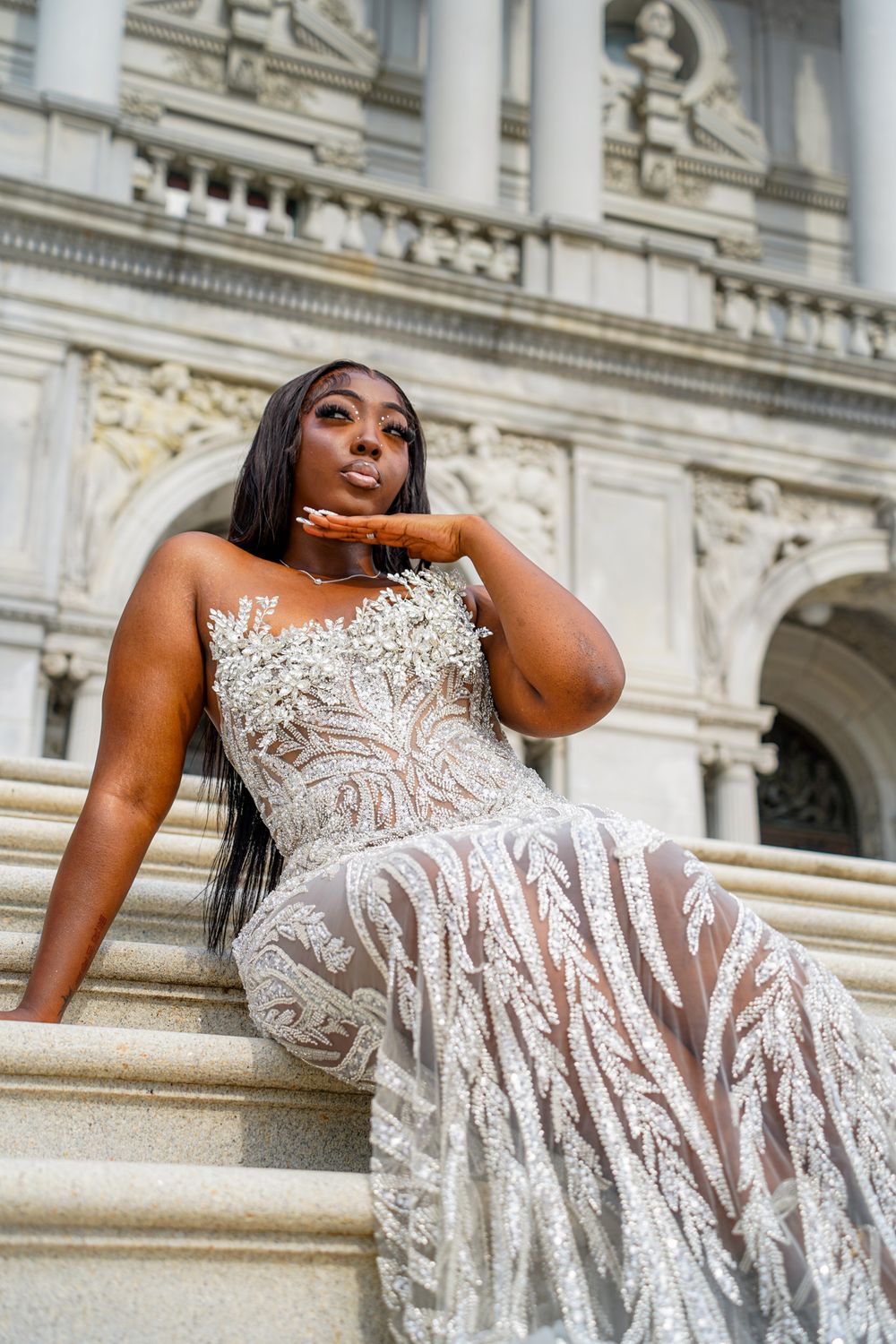A person in a white lace wedding dress poses elegantly on marble steps of a grand historic building.