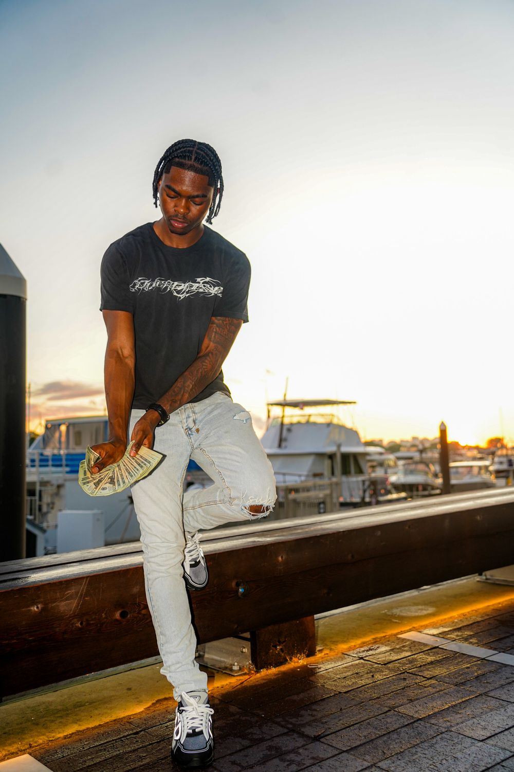 Person in black t-shirt and light jeans poses on dock with boats and sunset in background.