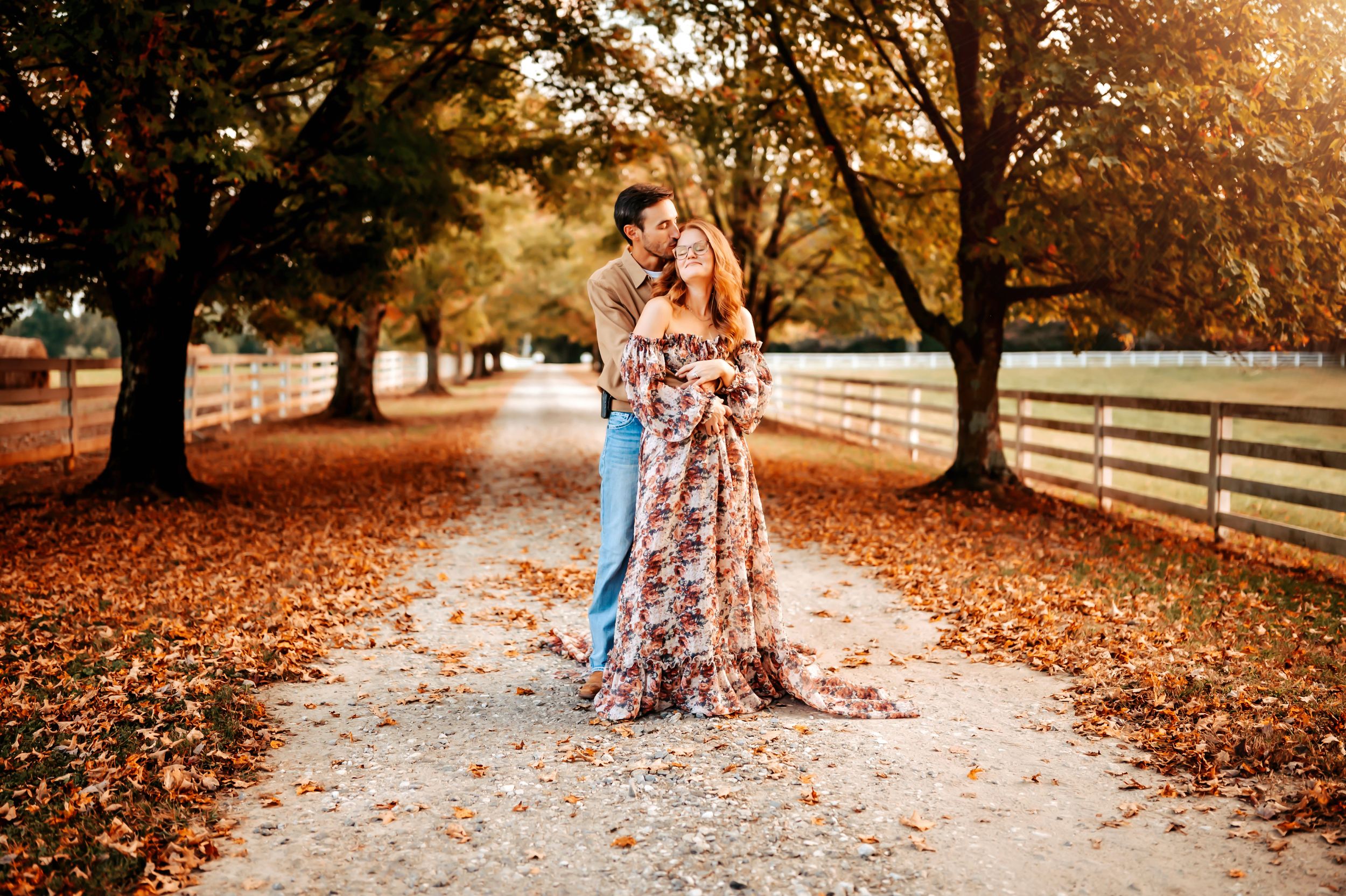 A couple embraces on an autumn path with fallen leaves and wooden fence during golden hour sunset.