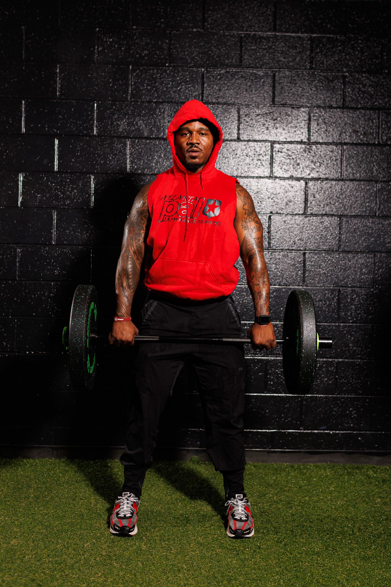 Athlete in red hoodie and black pants performs deadlift sequence against dark wall with artificial turf floor.