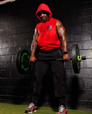 Athlete in red tank top performs dumbbell exercises against dark wall in gym setting.