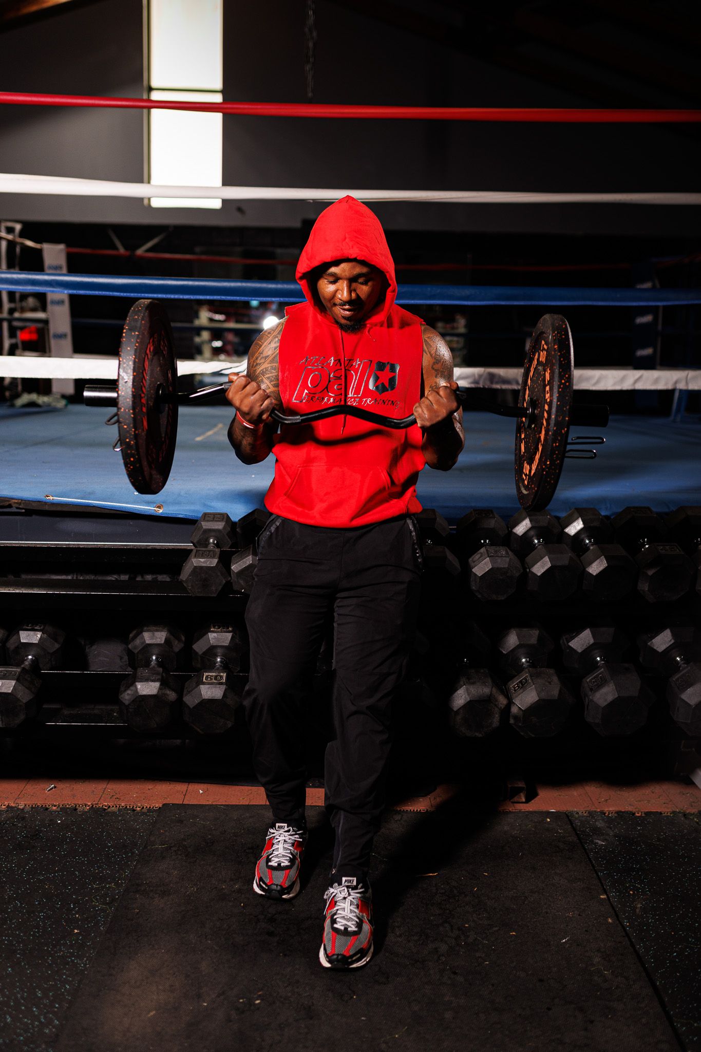 Person in red hoodie demonstrates weightlifting form with dumbbells in boxing gym environment.