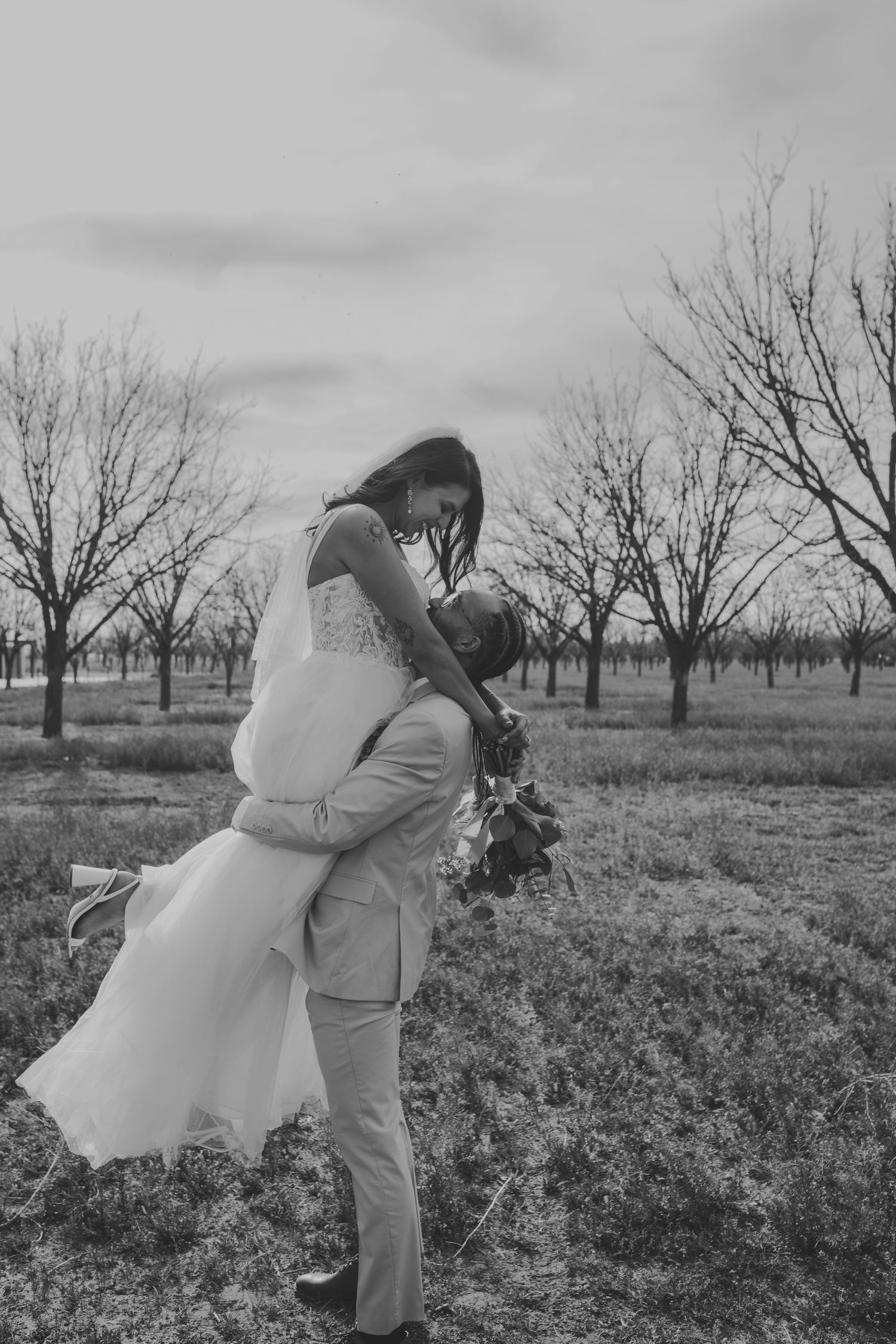 Black and white wedding photo of groom lifting bride among bare trees in an open field.