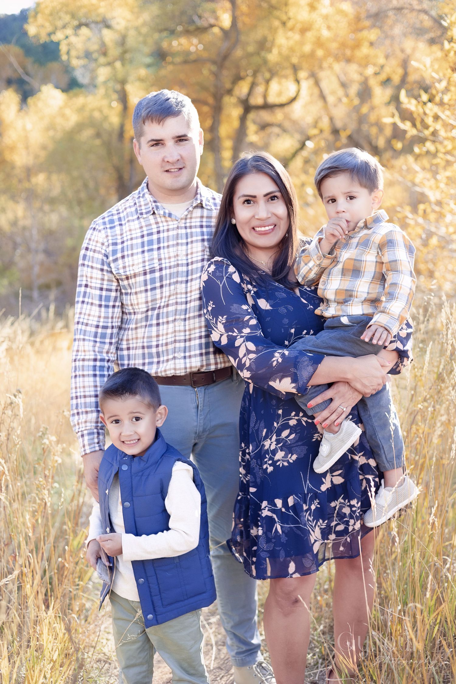 A family poses together in a field of tall grass during golden hour with autumn trees in the background.