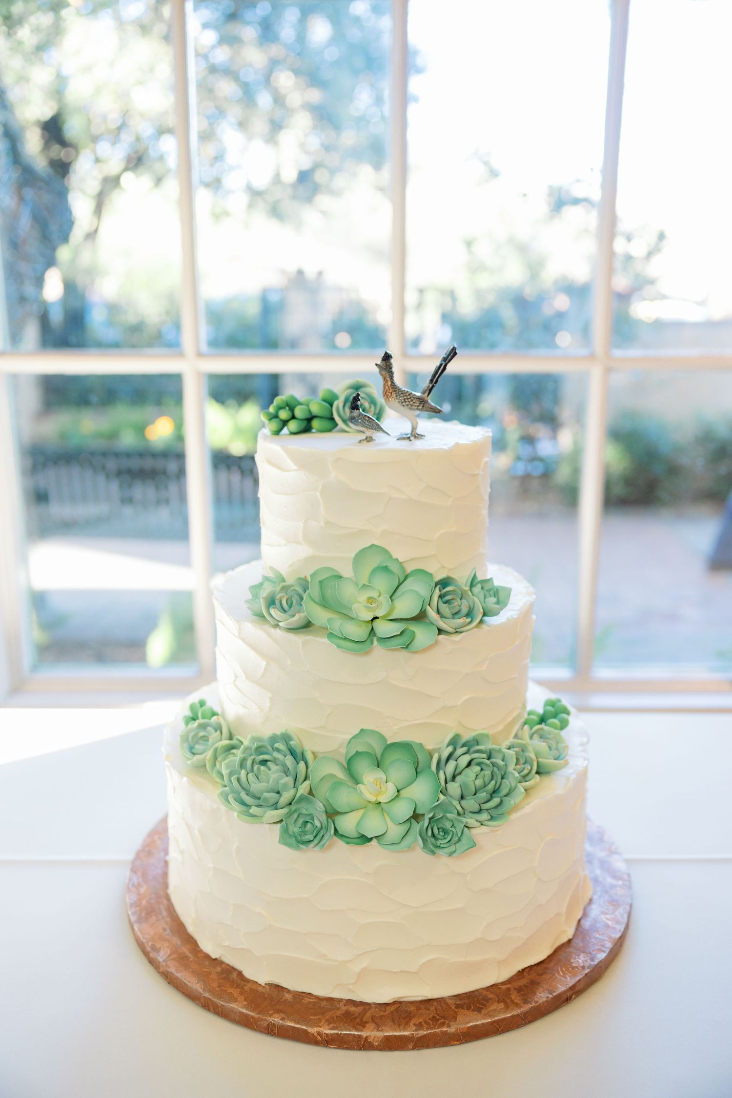 White two-tier wedding cake decorated with delicate mint green succulent designs on a wooden cake stand.