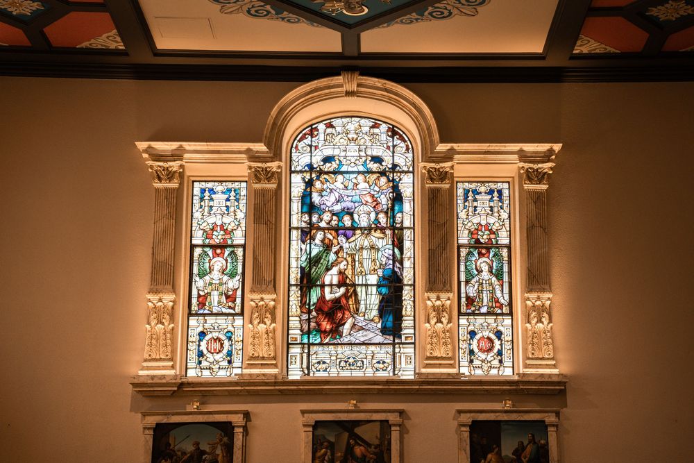 Ornate stained glass church window with religious artwork surrounded by decorative architectural framing in warm lighting.