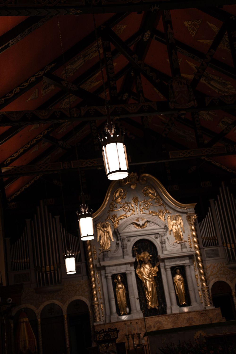 Ornate baroque church organ pipes and decorative carvings illuminated by hanging lanterns in a dimly lit cathedral interior.