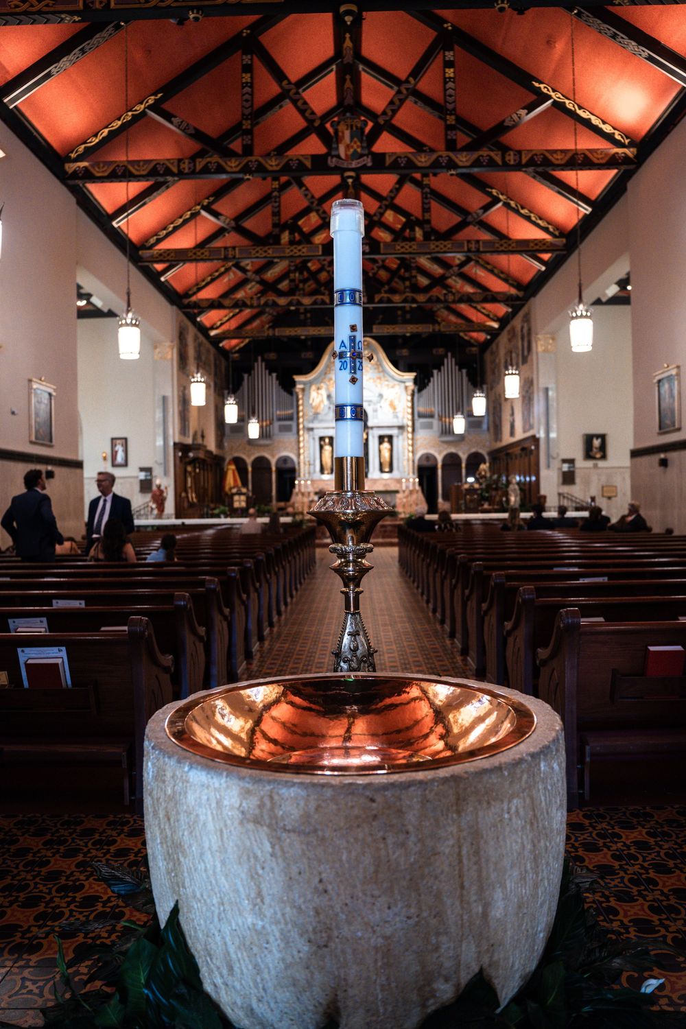 Interior of a church with exposed wooden beams, pendant lights, and a baptismal font in the foreground.