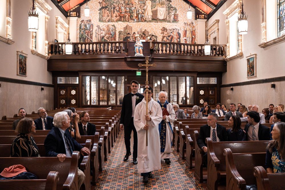 Wedding guests sit in wooden pews inside a historic church with ornate architectural details and high ceilings.