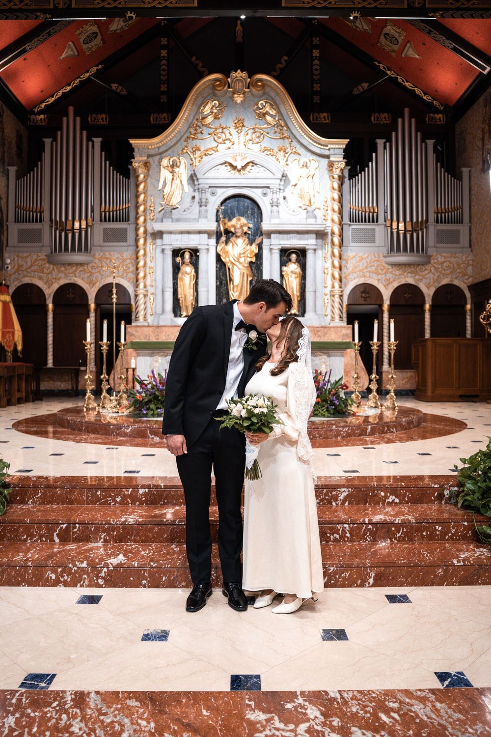 A couple shares an intimate moment during their wedding ceremony in an ornate church with marble steps and pipe organs.