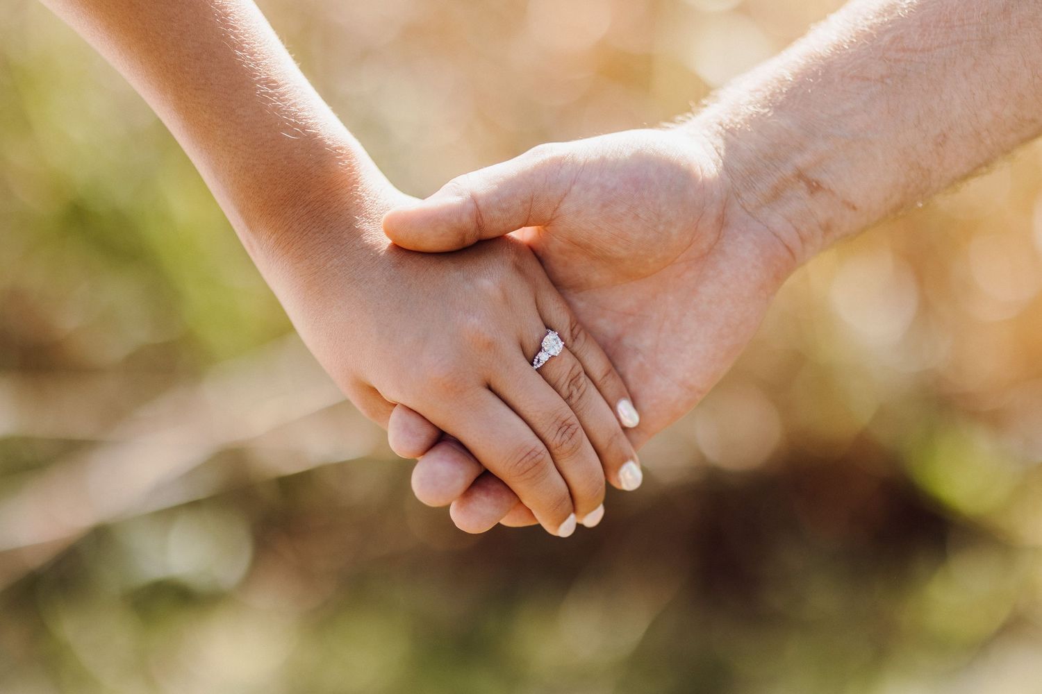 Close-up of two hands holding each other with a sparkling engagement ring in soft natural light.