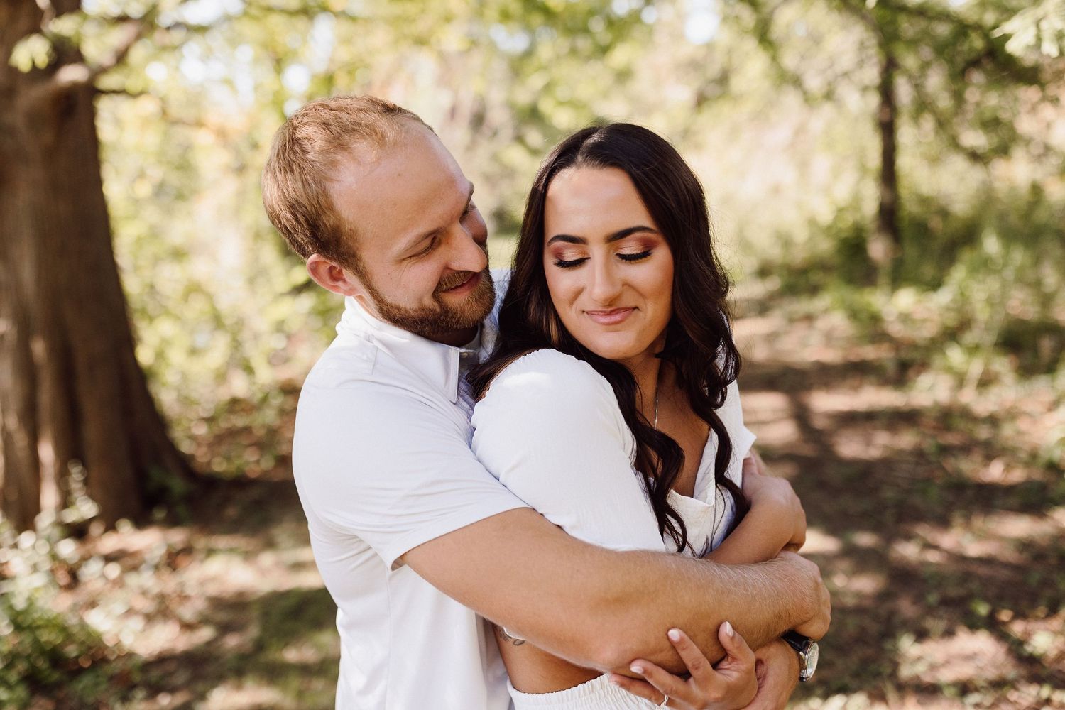 A couple shares an intimate embrace while standing together in a wooded outdoor setting.