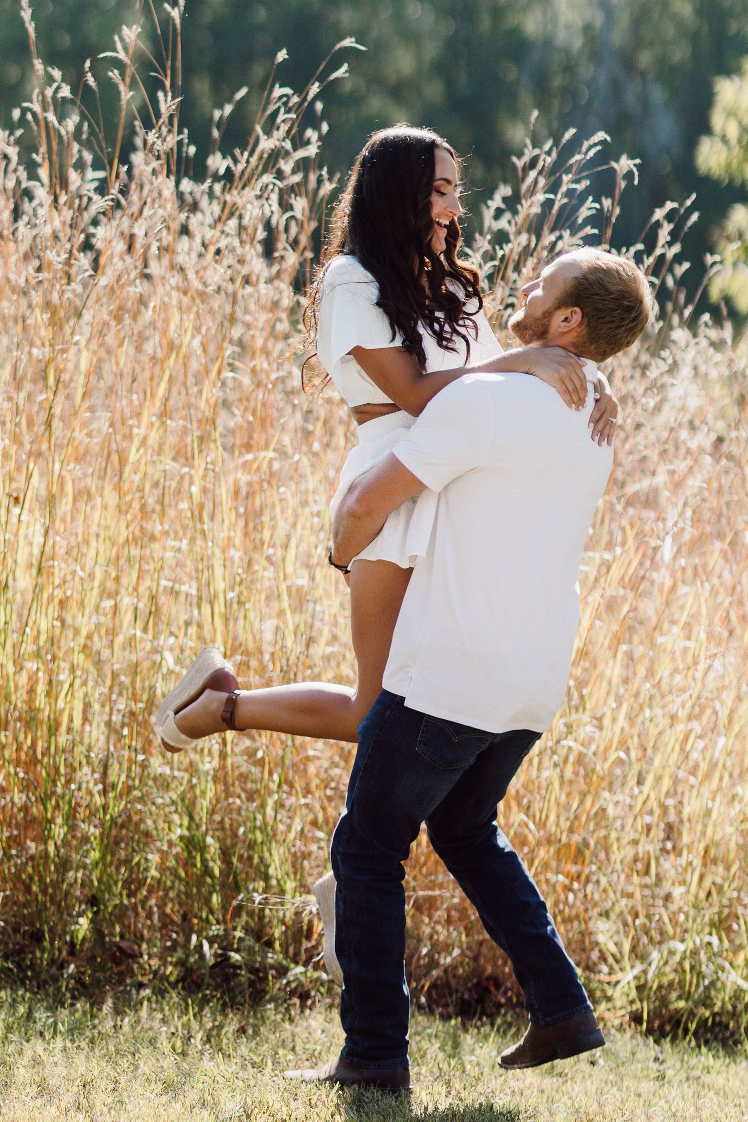 A couple in white outfits embraces playfully in a field of tall golden grass on a sunny afternoon.
