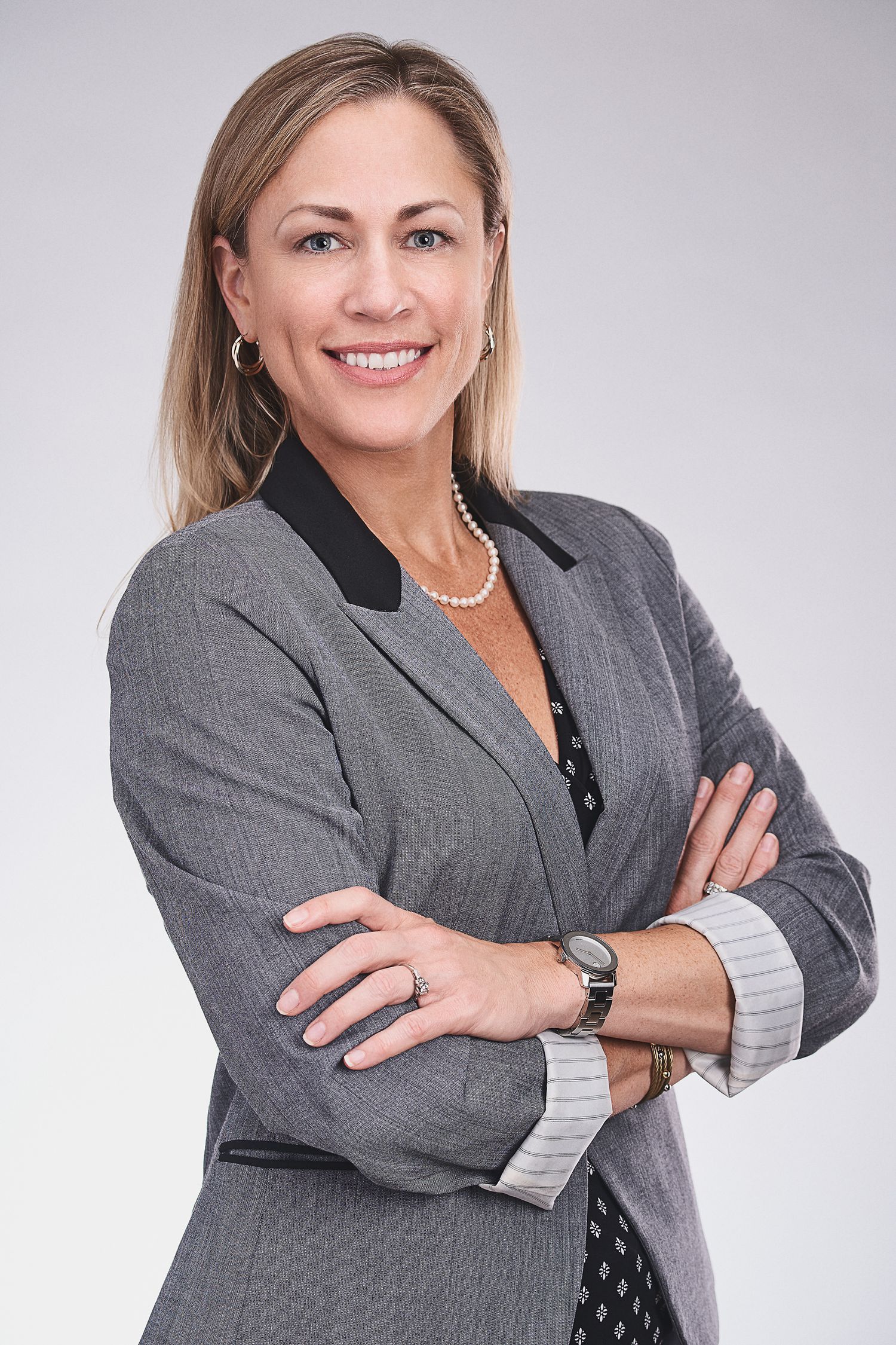 Professional businesswoman in gray blazer smiling with arms crossed against light background.