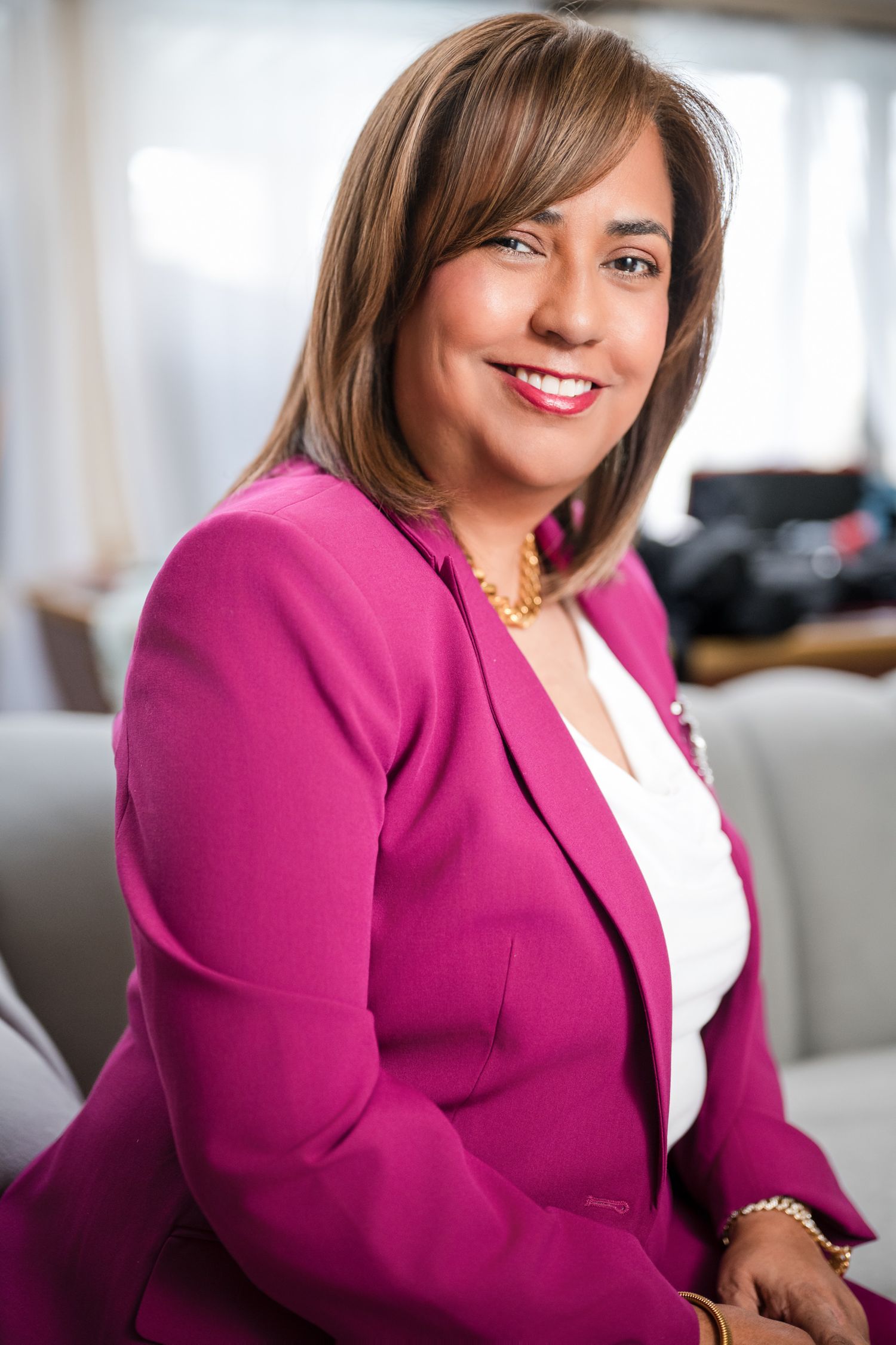 Smiling businesswoman in pink blazer seated in bright office wearing gold necklace.