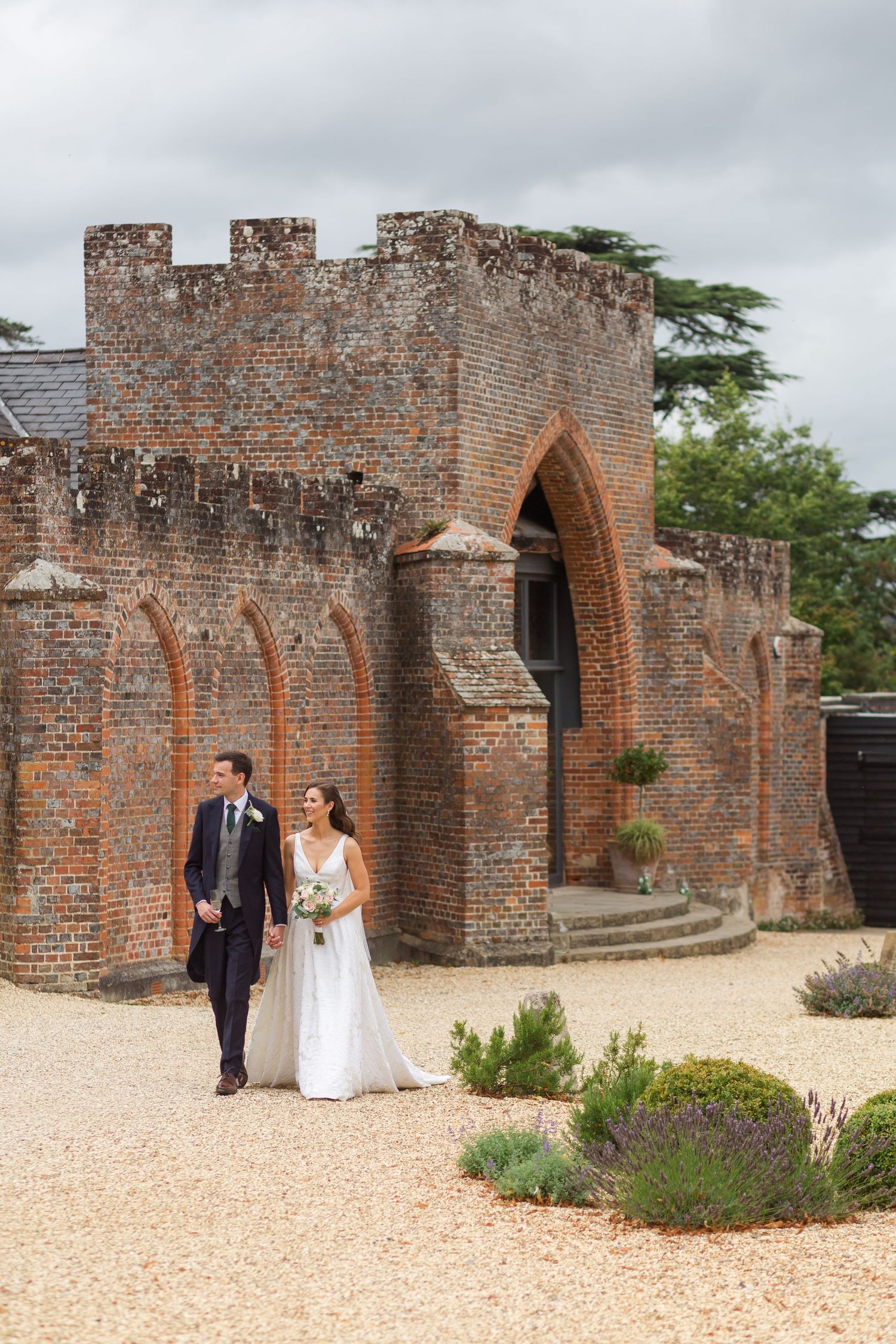 A couple walks along the rustic brick walls and gravel path at a historic venue during their wedding day.