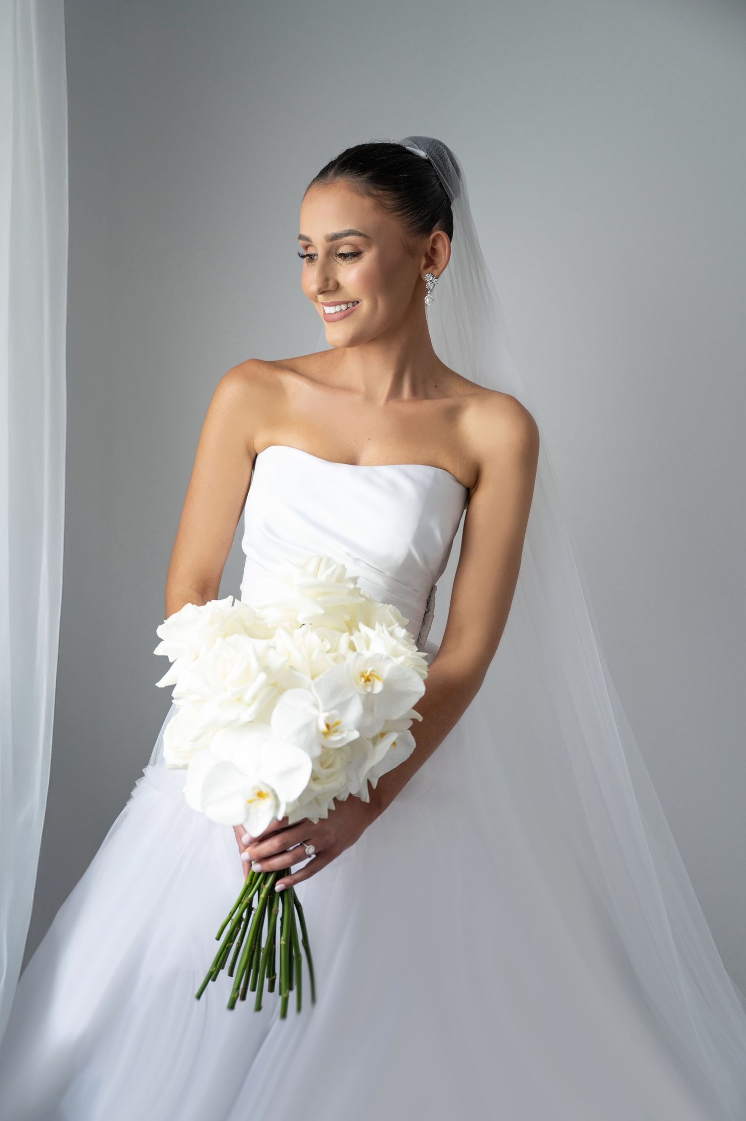 Elegant bride in strapless white gown holds bouquet of white orchids with veil and updo hairstyle.