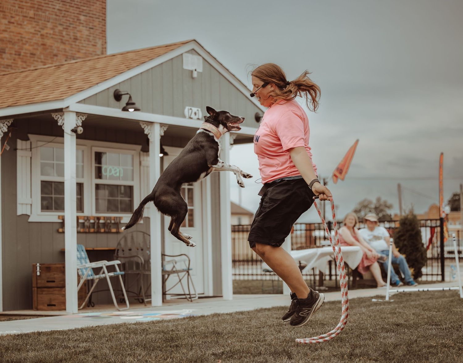 A woman in a pink shirt jumps rope with an energetic black dog at an outdoor event.