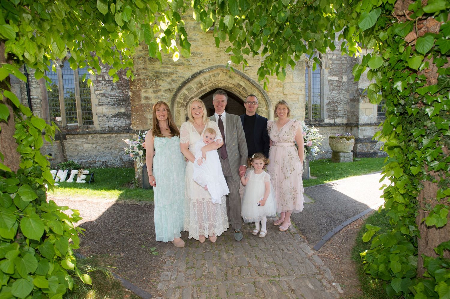 A family is gathered in front of a church with a tree behind, celebrating a christening, photographed by Abigail's Studio in Buckinghamshire
