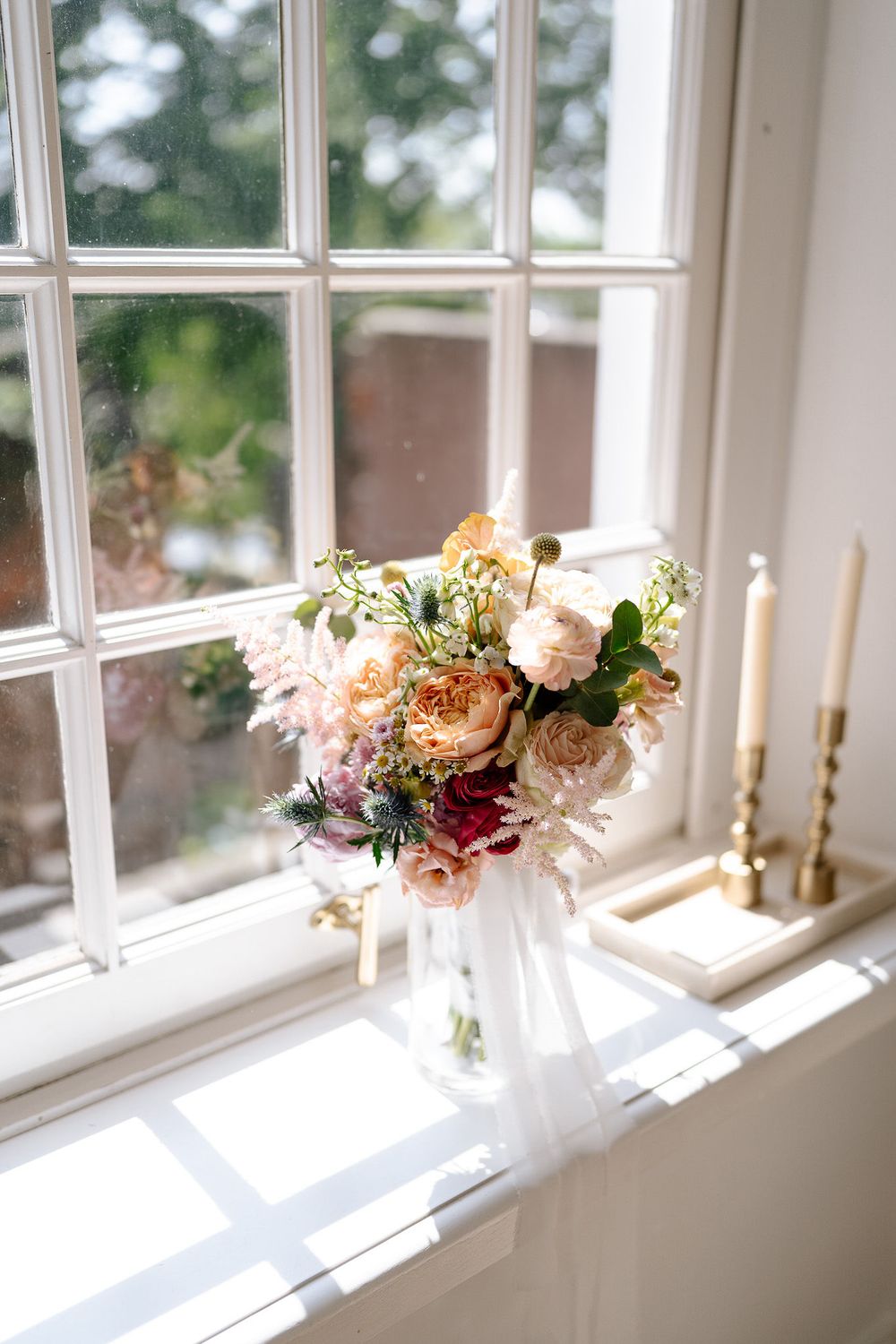 Sunlit windowsill decorated with elegant floral arrangements and golden candlesticks in a bright white interior space.