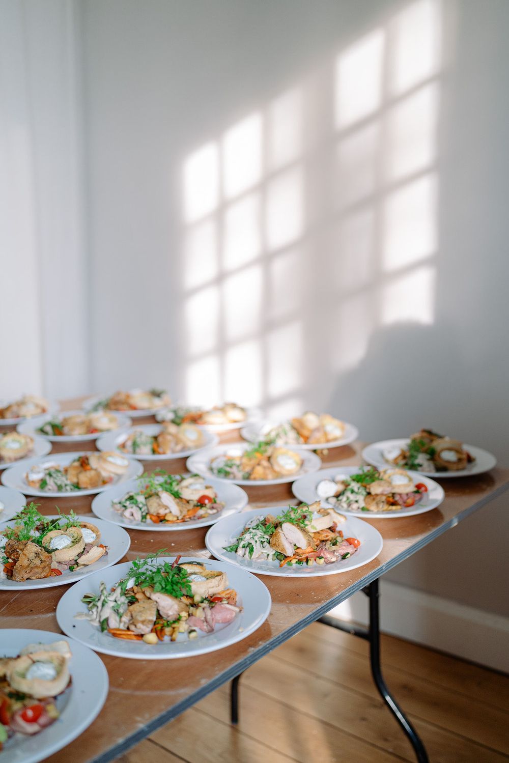 Multiple plates of elegantly plated meals arranged on a folding table with sunlight casting window shadows on the wall.