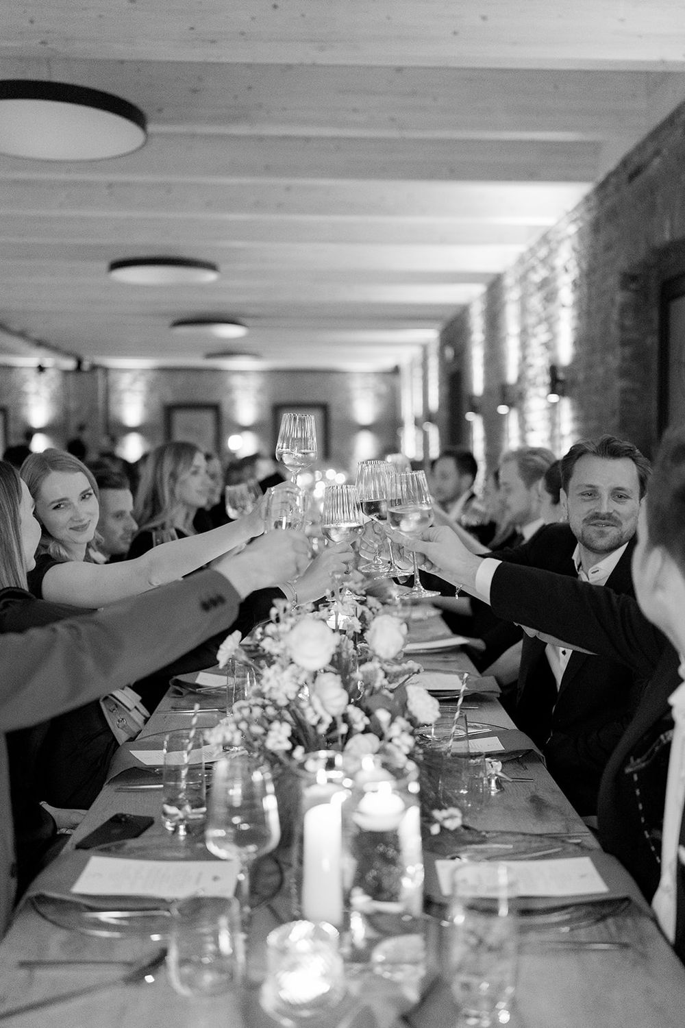 Wedding guests raise champagne glasses in a toast at a long candlelit reception table.