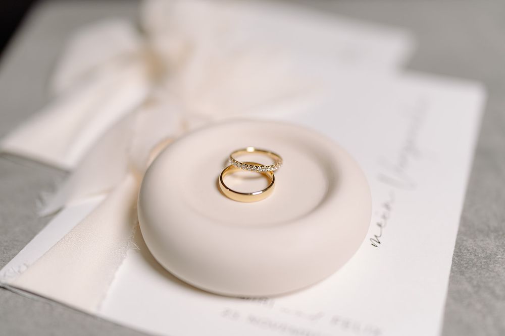 A delicate gold wedding ring displayed on a small white ceramic ring holder dish against a light background.