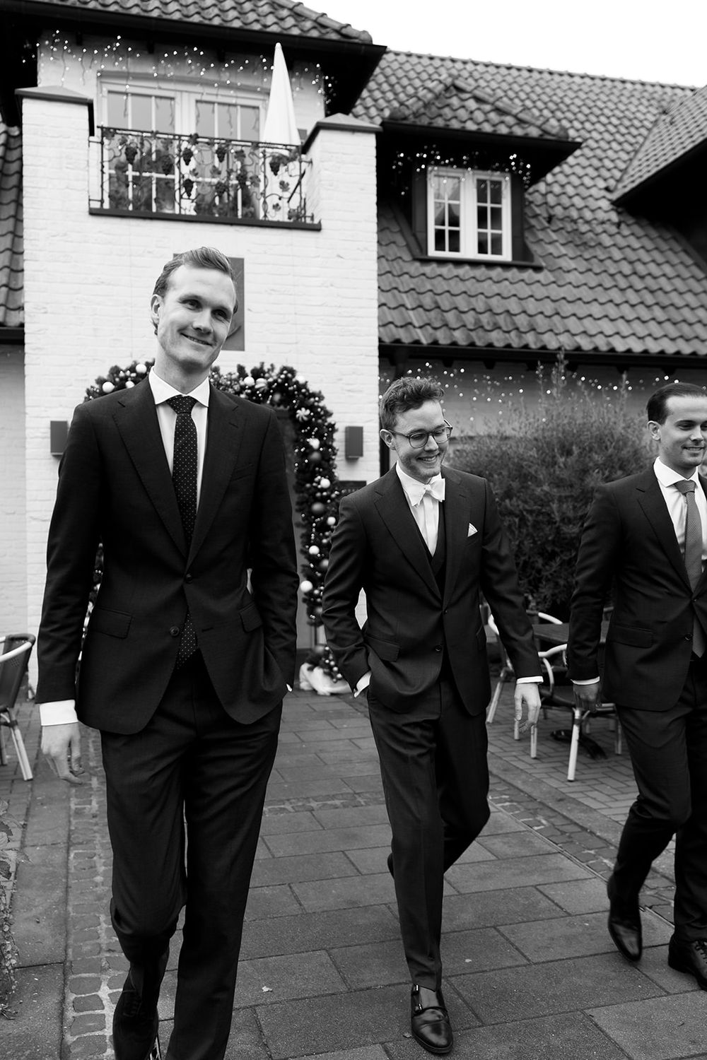 Three men in formal black suits and ties walking together outside a house with a tiled roof and white walls.