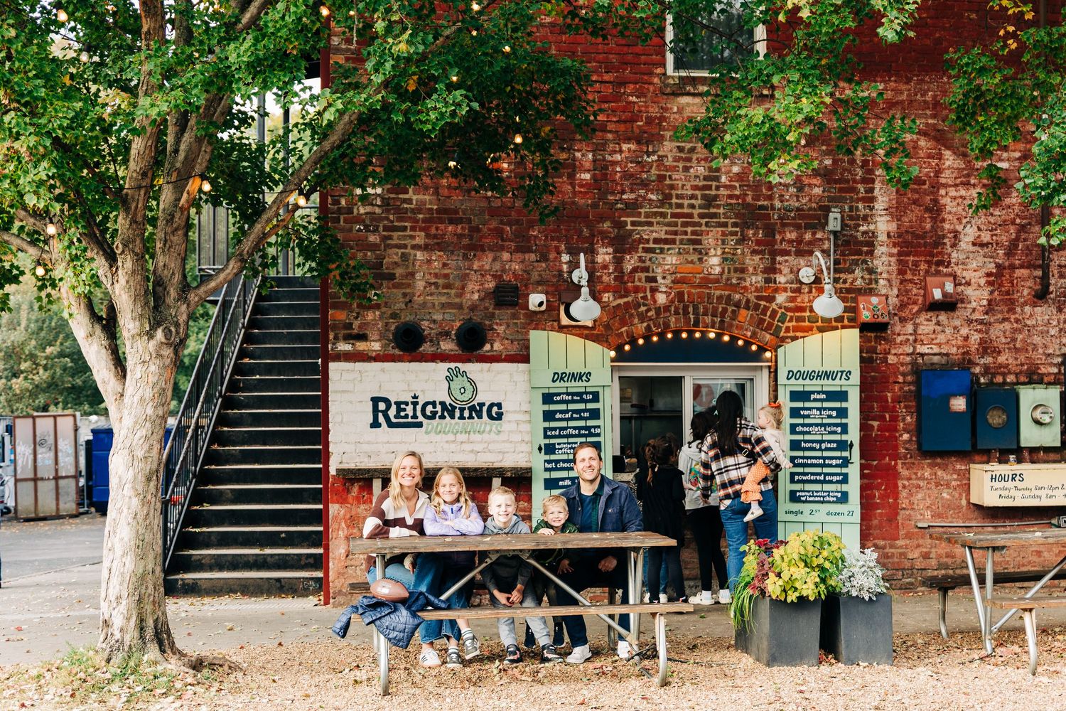 People gather at outdoor tables in front of a rustic brick building waiting on doughnuts and green shutters.