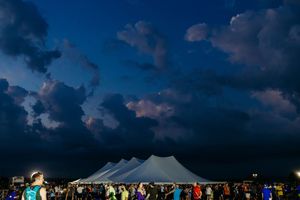A large white event tent illuminated at dusk with dramatic dark blue clouds and people gathered outside.