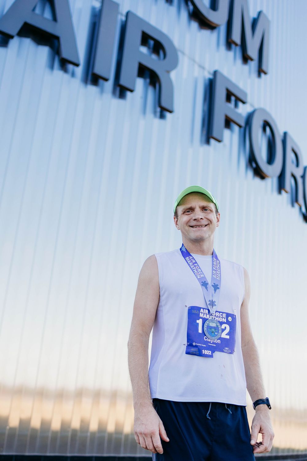 Runner in white tank top and green cap smiling during outdoor training session at sunset.