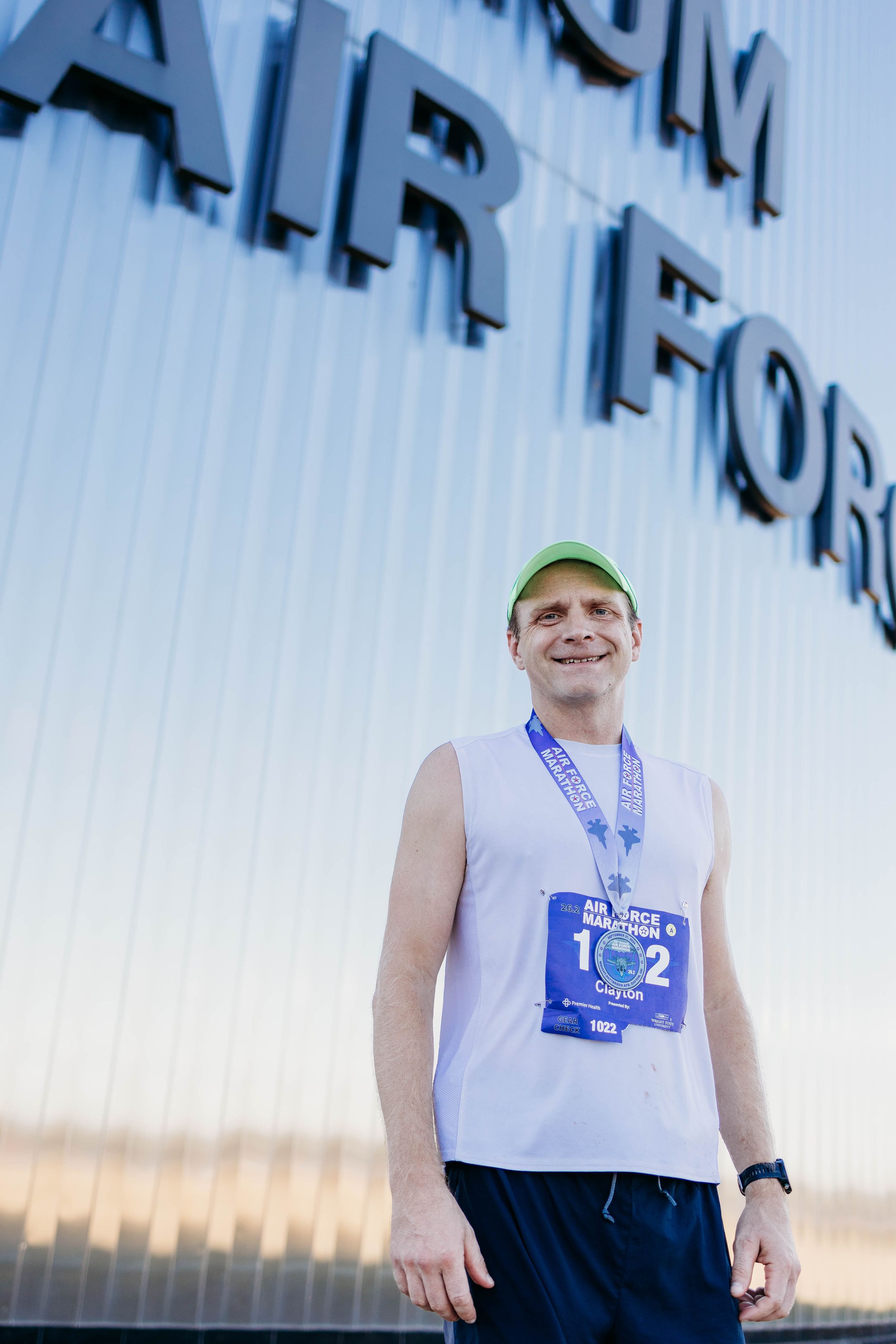 Runner in white tank top and green cap smiling during outdoor training session at sunset.